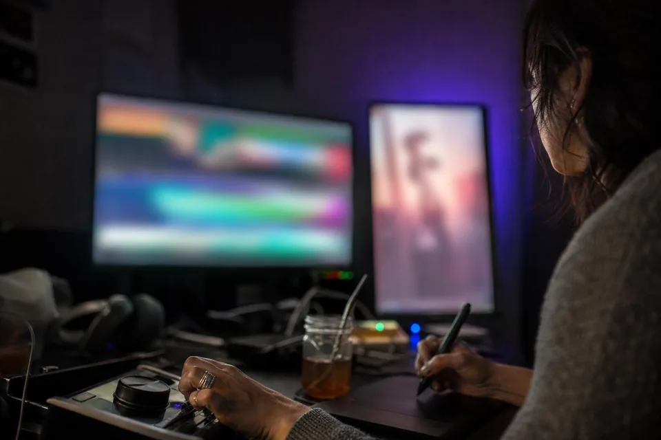 A video editor at night marking cut points on a pen tablet, with a wide timeline monitor and a second monitor turned upright for a vertical clip preview blurred in the background
