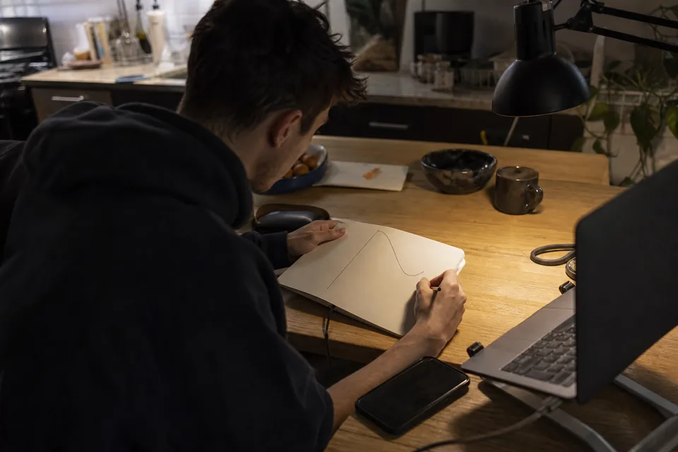 A creator sketching a curve in a notebook at a kitchen table at night