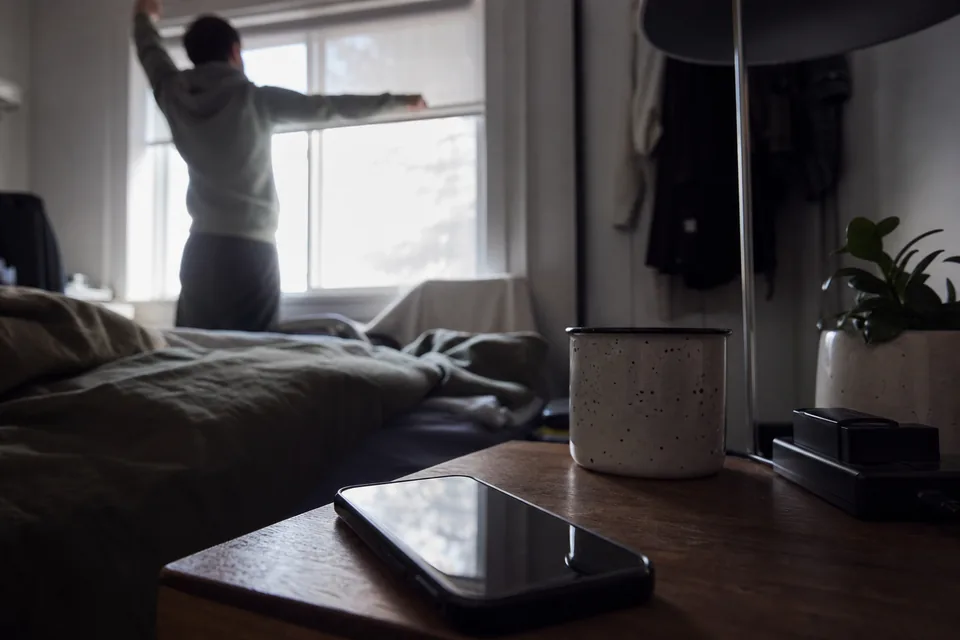 A phone on a nightstand at dawn next to coffee and a charging camera battery
