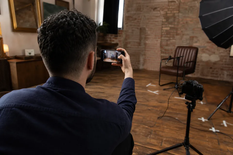A creator checking the framing on a phone in front of an empty chair and microphone