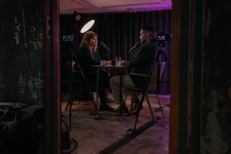Two podcast hosts across a table in a dim studio, cameras on tripods pointed at each of them